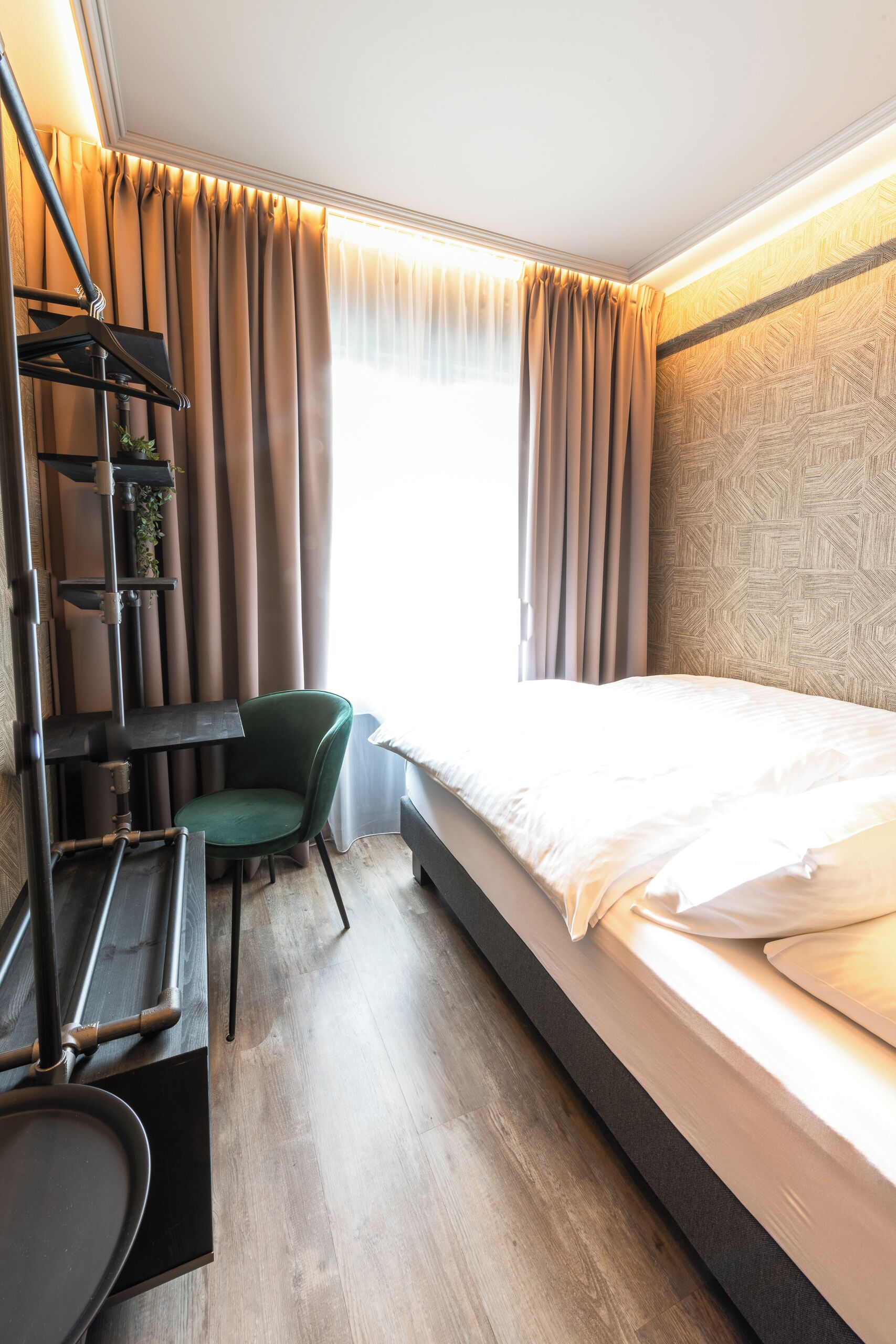Een hotelkamer met een bed, een bureau met stoel, een plank met planten en een raam met gordijnen.