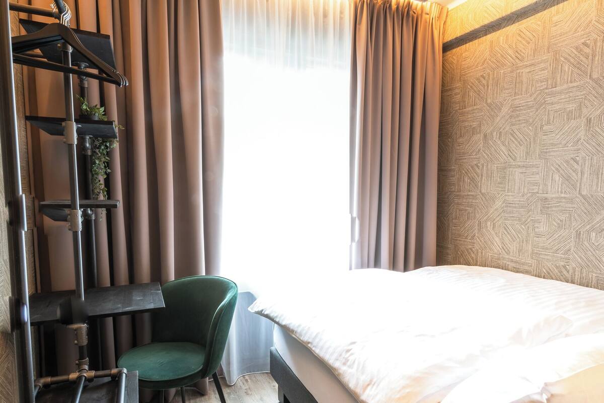 Una habitación de hotel con cama, escritorio y silla, estante con plantas y ventana con cortinas.