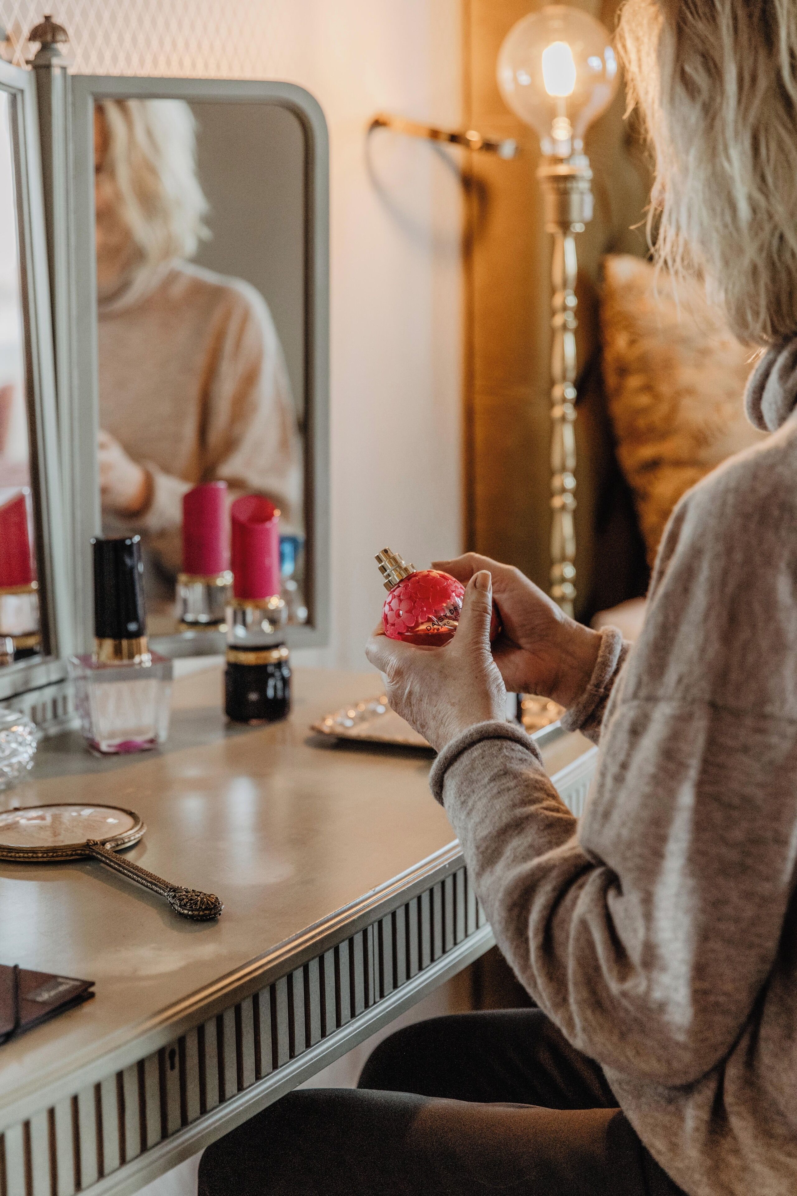 Una persona sostiene un objeto rojo en un tocador con espejo, una lámpara y varios artículos de cosméticos sobre la encimera.