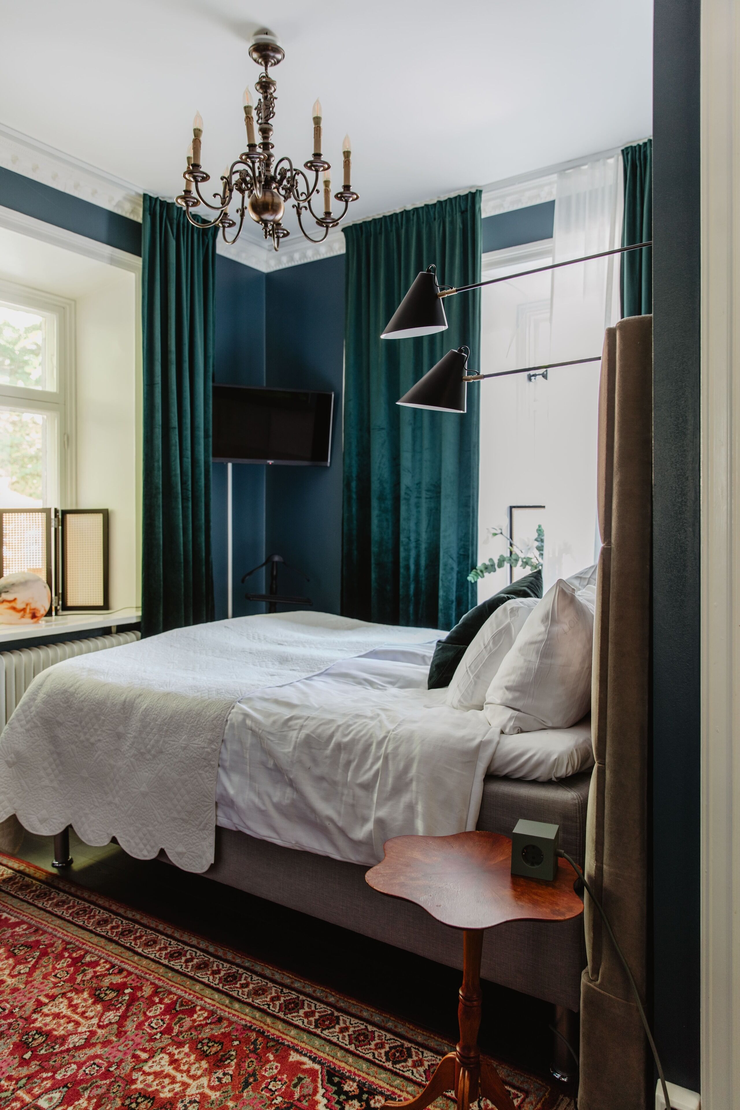 A bedroom with a bed, a desk, a chair, a TV, and a chandelier.