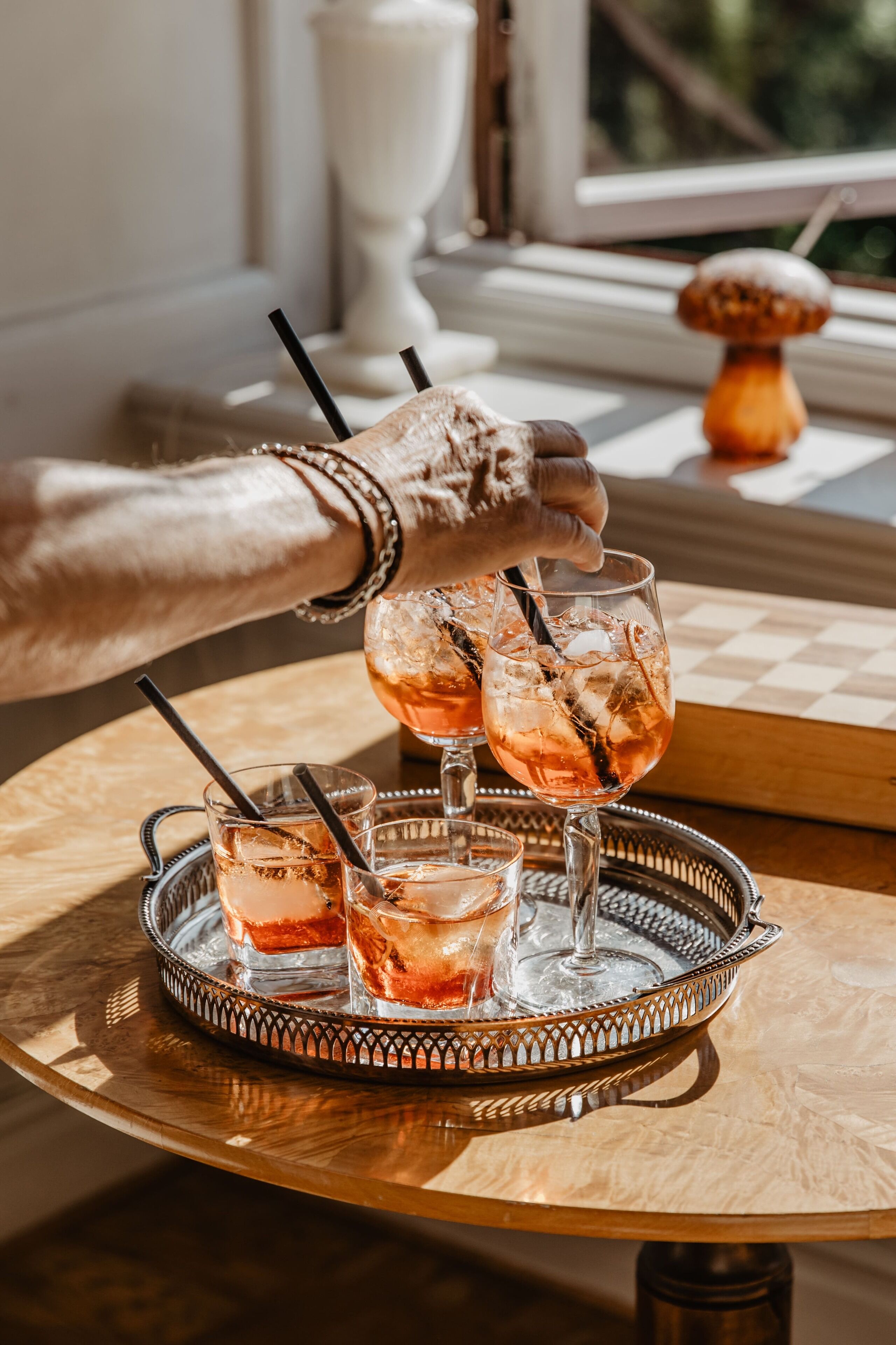Un plateau avec trois verres contenant une boisson rose, une main tenant un verre, et un jeu d’échecs en arrière-plan.