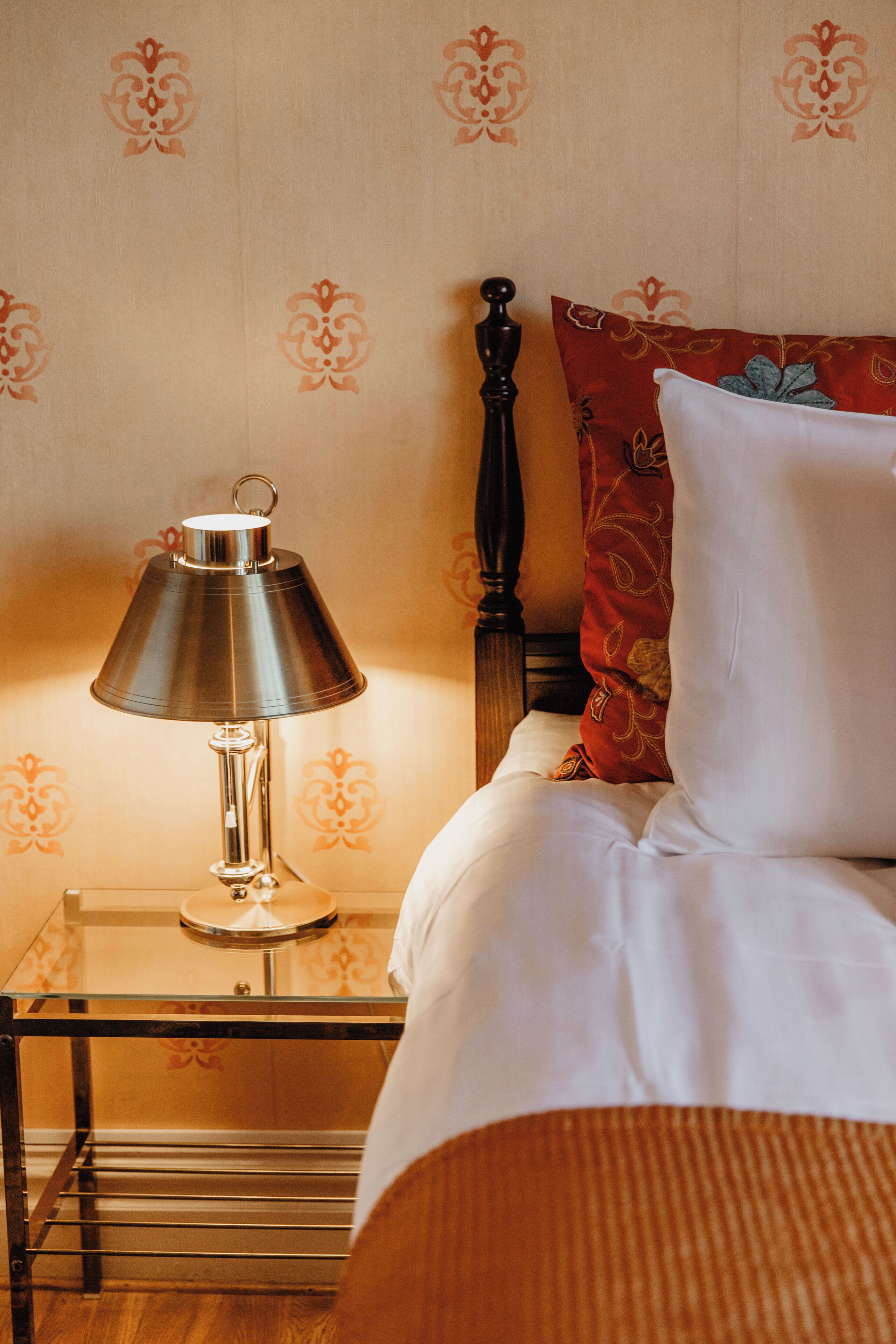 A neatly made bed with white linens, a red patterned pillow, and a metal lamp on a bedside table.
