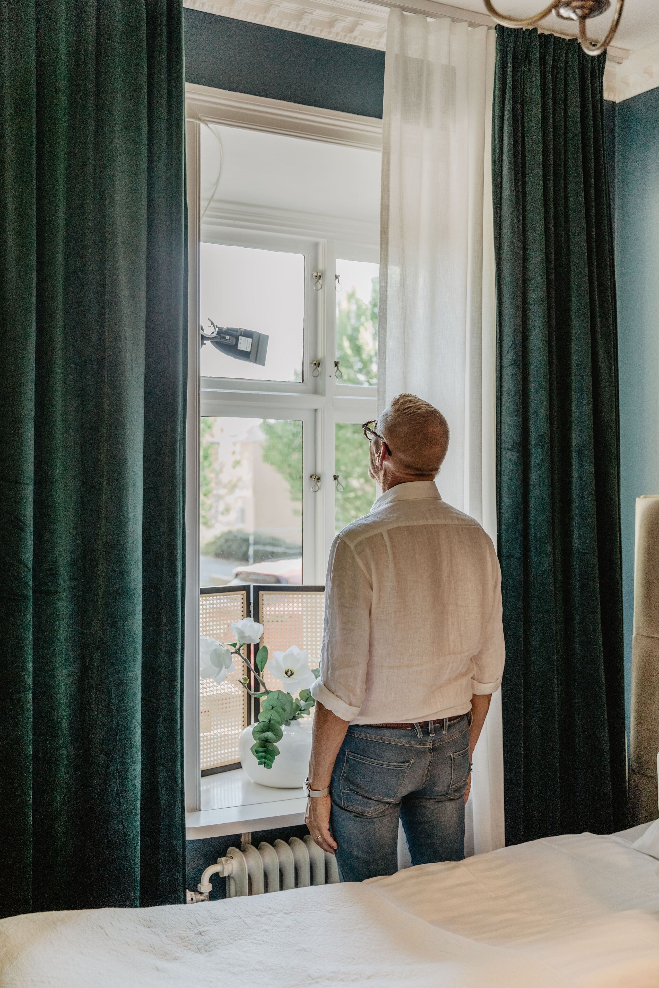 A man standing by a window in a room with a bed, a radiator, and a chair.