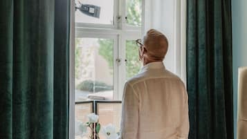 A man standing by a window in a room with a bed, a radiator, and a chair.