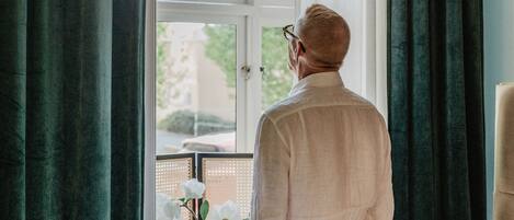 A man standing by a window in a room with a bed, a radiator, and a chair.