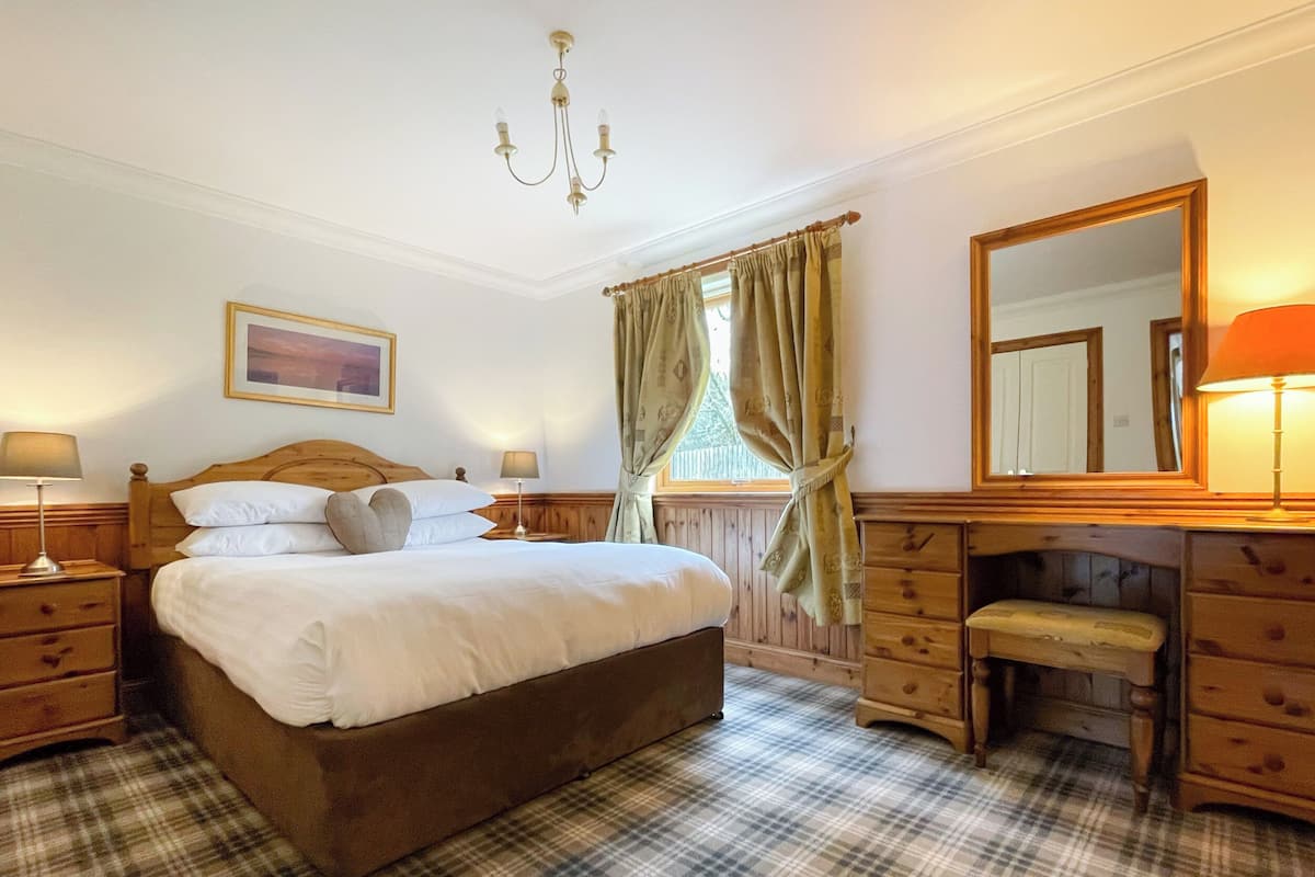 A bedroom with a large bed, a wooden dresser, a chair, a nightstand, a lamp, a mirror, and a window with curtains.
