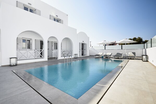 Un área de piscina moderna con una piscina azul cristalina, arquitectura blanca y tumbonas.