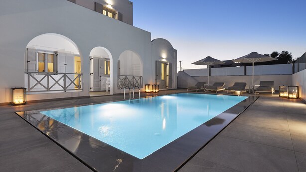Un área de piscina moderna con una piscina rectangular, sillas de descanso y un edificio con arcos y luces.