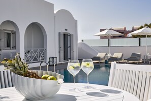 Un ambiente junto a la piscina con una mesa blanca, dos vasos de una bebida transparente, un cuenco con frutas y un edificio blanco con arcos al fondo.
