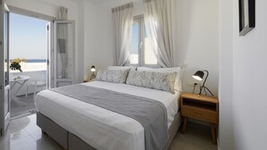 Un dormitorio con cama, mesitas de noche, una ventana con cortinas, una puerta que da a un balcón y una pequeña mesa con una silla.