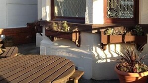 Un espacio al aire libre con mesas y bancos de madera, un edificio blanco con una ventana y una planta en maceta.