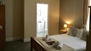 Habitación de hotel con cama, mesita de noche, lámpara y vista a un baño por una puerta abierta.