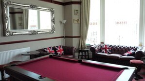Una mesa de billar en una habitación con un espejo grande, dos sofás con banderas británicas y una ventana con cortinas.