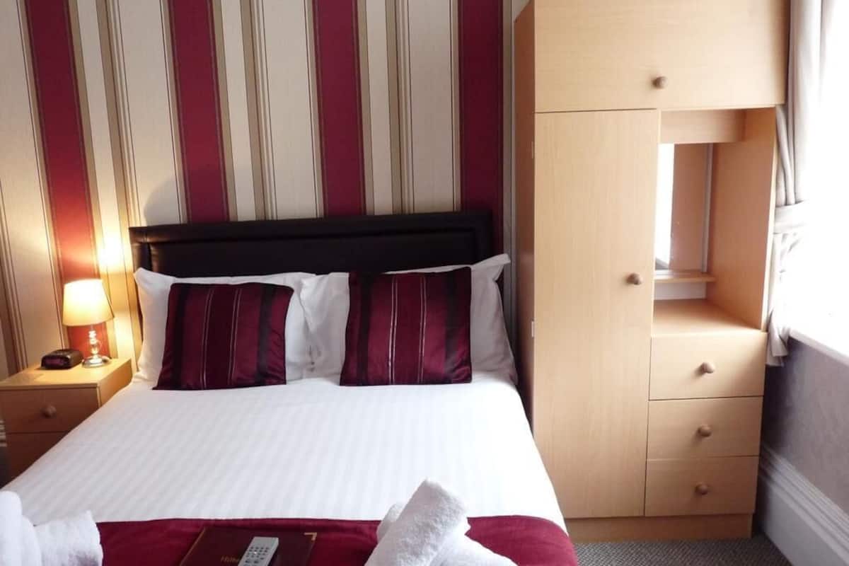 Habitación de hotel con cama, mesita de noche con lámpara, armario de madera y ventana con cortinas.