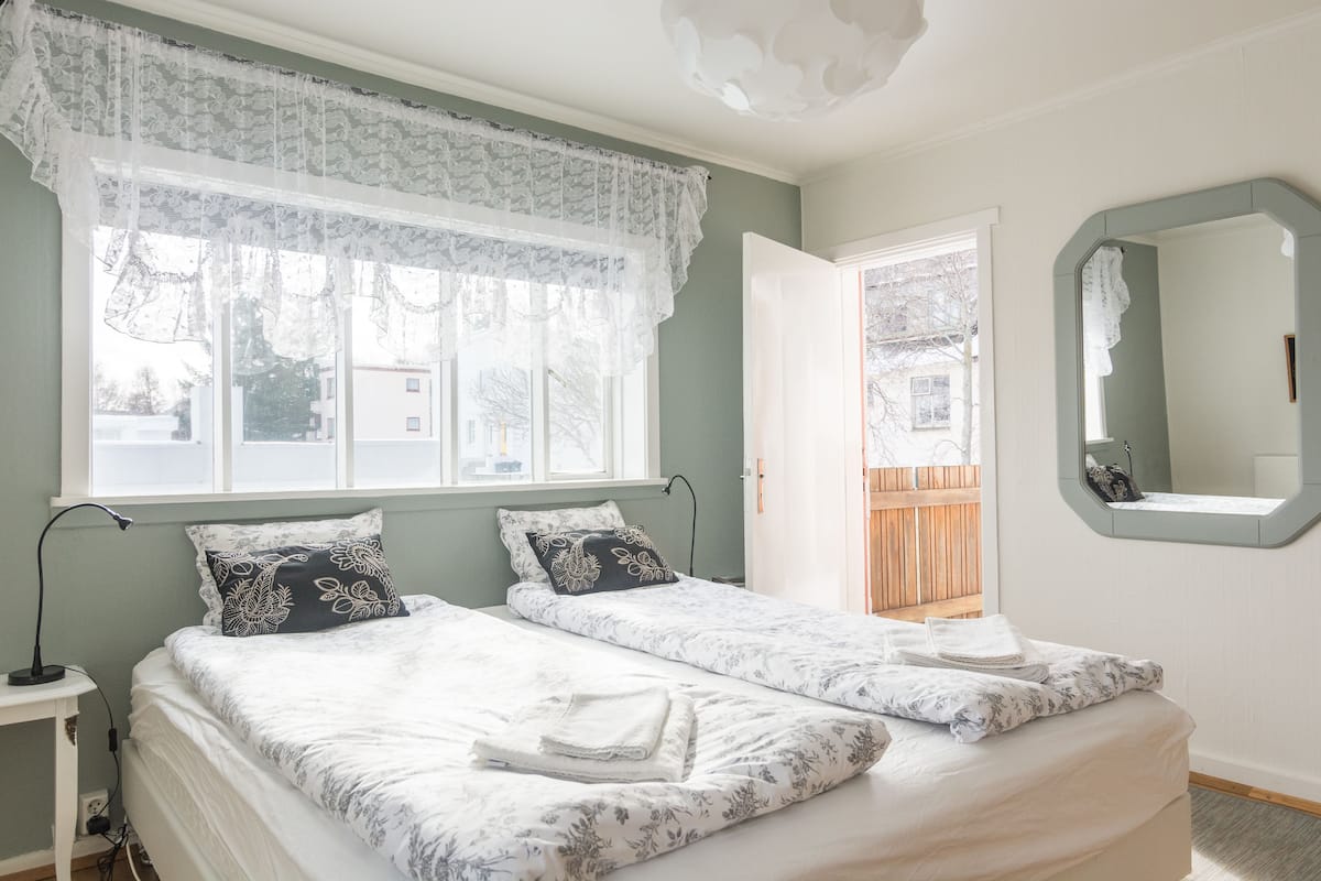 A bedroom with two beds, a window with lace curtains, a bedside table with a lamp, and a mirror on the wall.