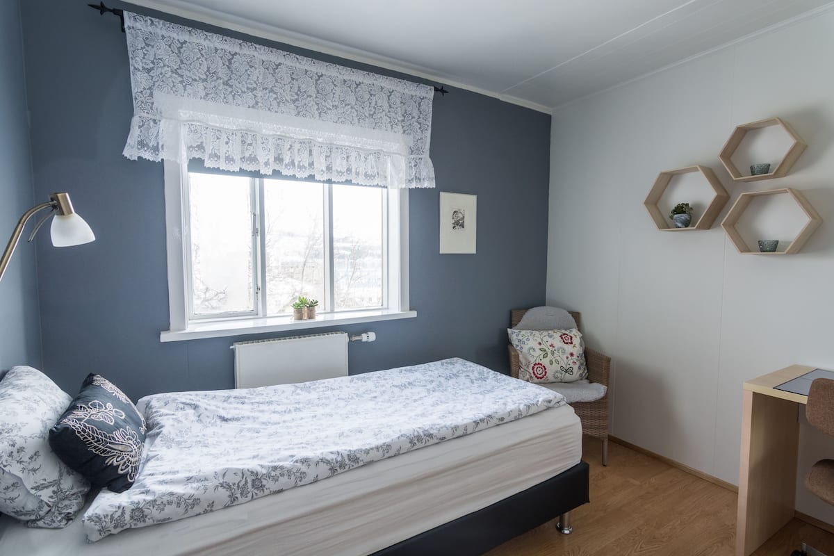 A bedroom with a bed, a window with lace curtains, a desk, and hexagonal shelves with plants.
