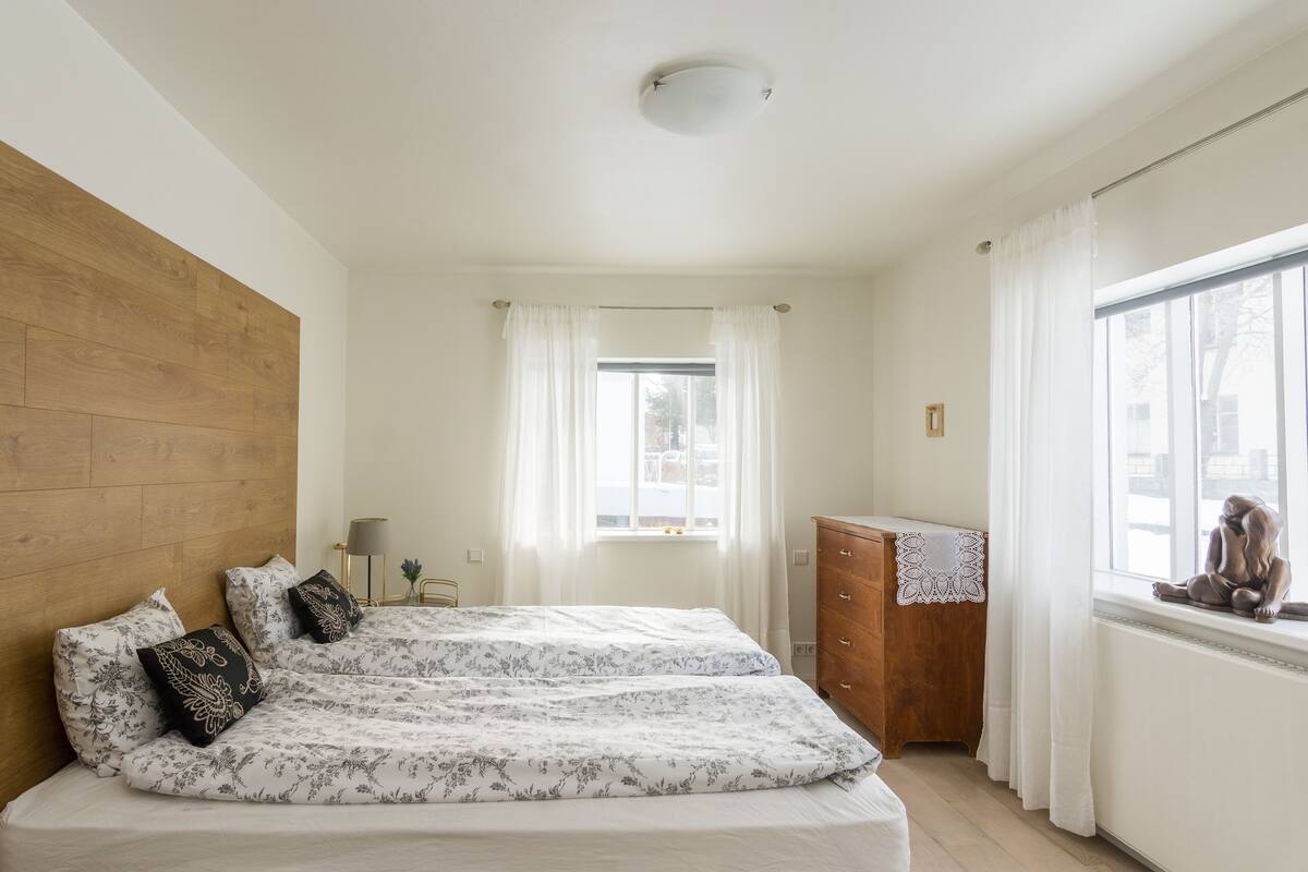 A bedroom with two beds, a wooden headboard, a dresser, a window with curtains, and a small statue on the windowsill.