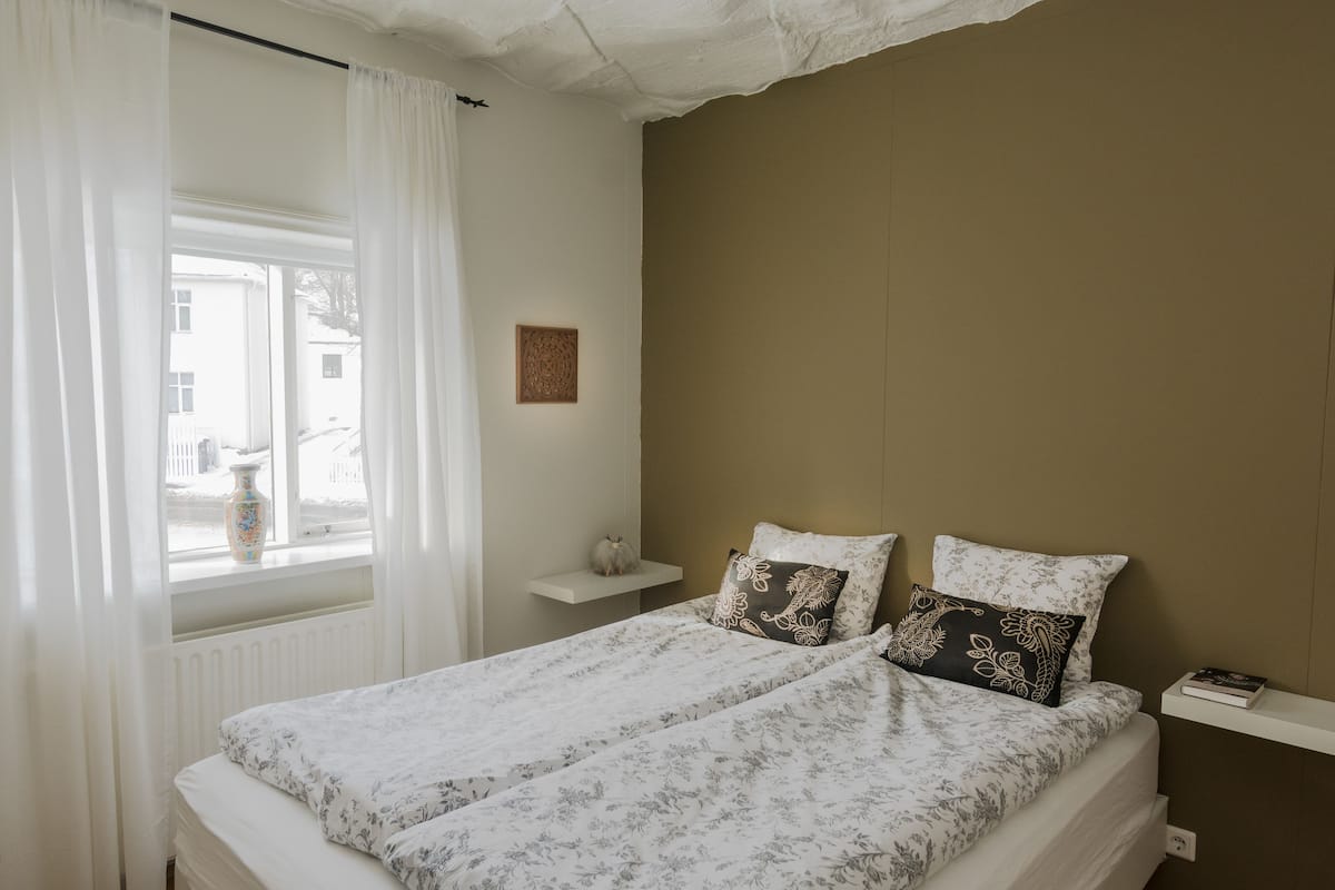 A bedroom with a bed, a window with curtains, a small shelf, and a wall-mounted picture.