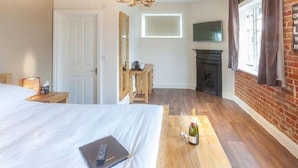 Un dormitorio con cama, mesita de noche con lámpara, escritorio de madera, chimenea y televisor montado en la pared.