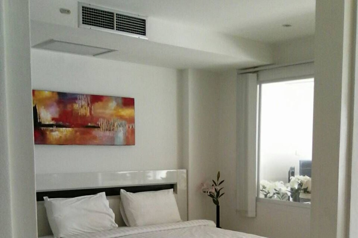 A modern hotel bedroom with a large bed, a painting on the wall, and a window with a view.