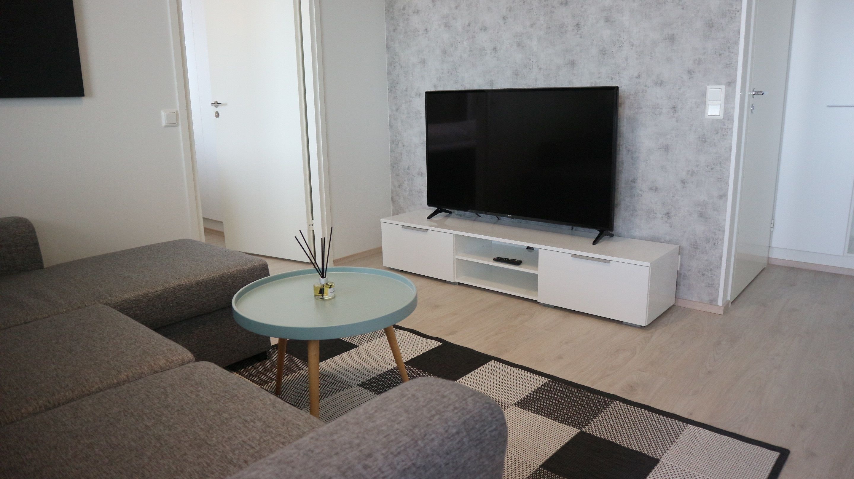 Een moderne woonkamer met een flatscreen televisie op een witte tv-kast, grijze banken, een kleine ronde tafel en een tapijt met een patroon.