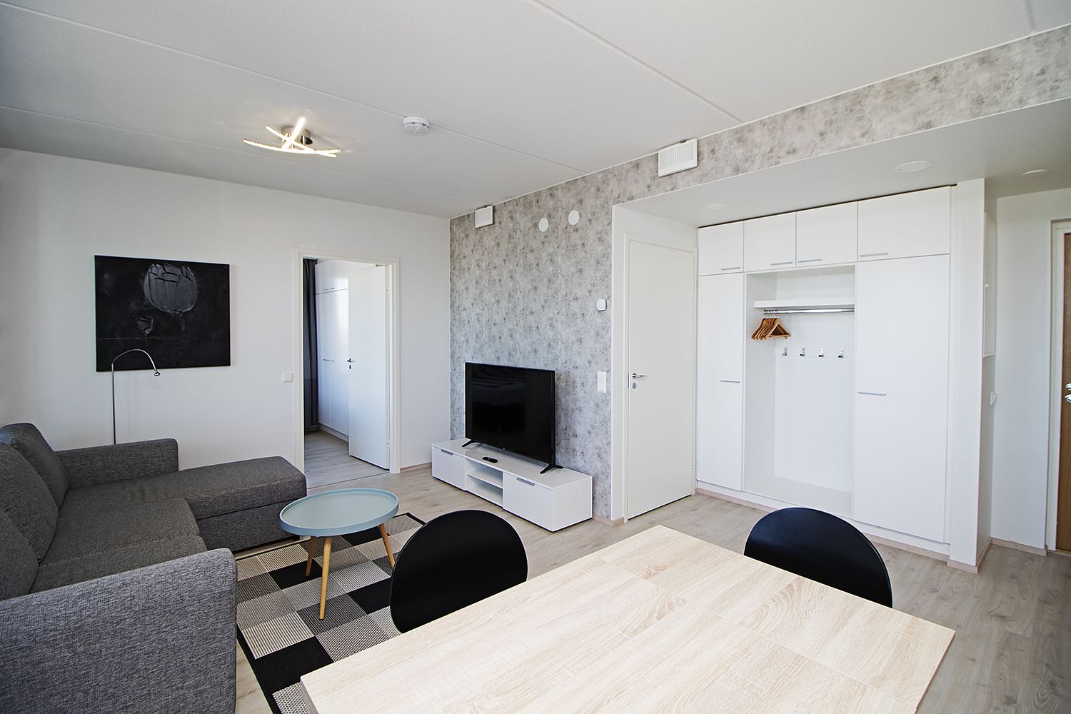 A modern living room with a grey sofa, a TV on a white stand, a small round table, and a wooden dining table.