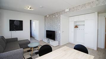 A modern living room with a grey sofa, a TV on a white stand, a small round table, and a wooden dining table.