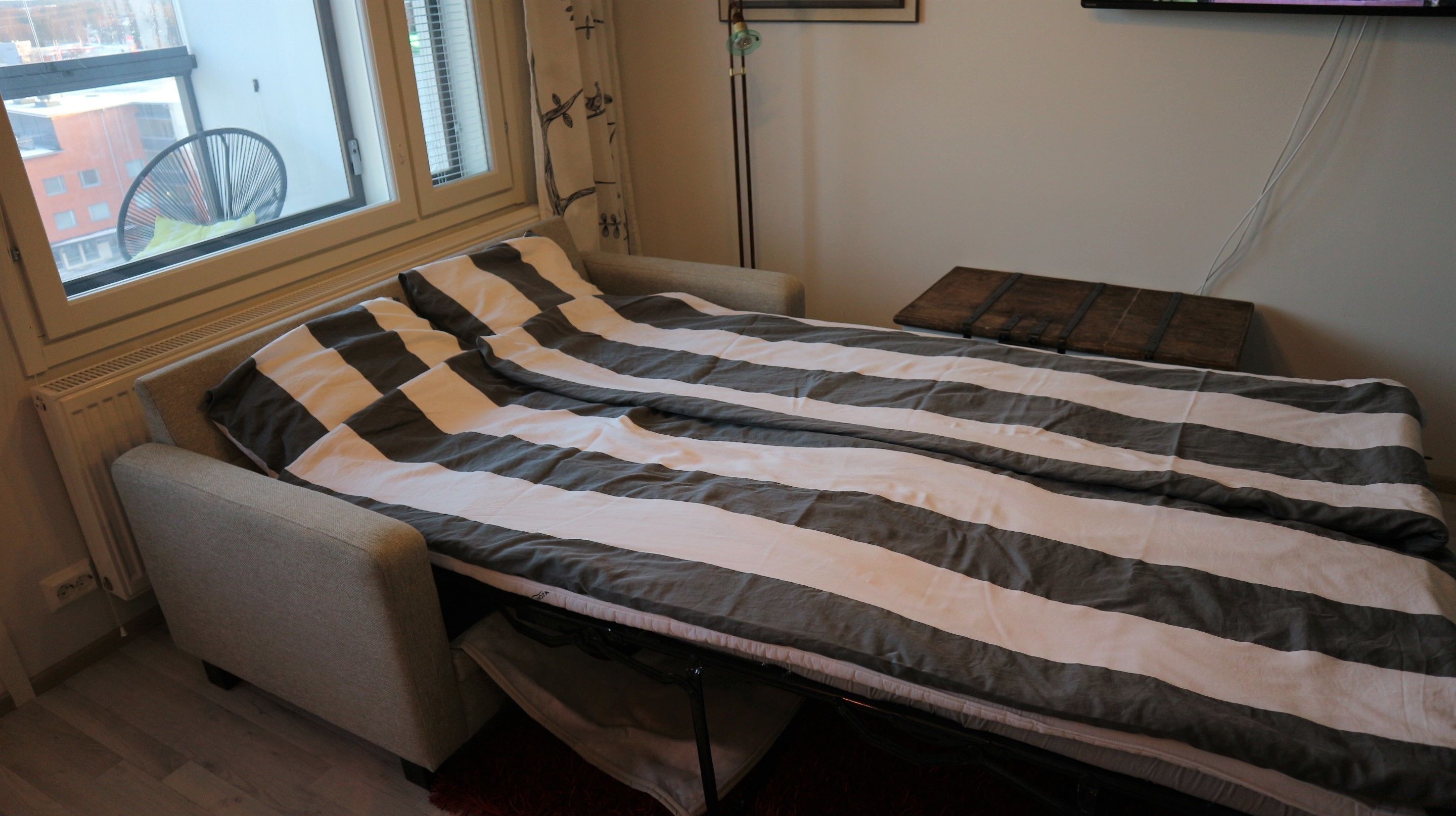 A compact bed with a striped blanket in a room with a radiator, a chair, and a window with a view of buildings.