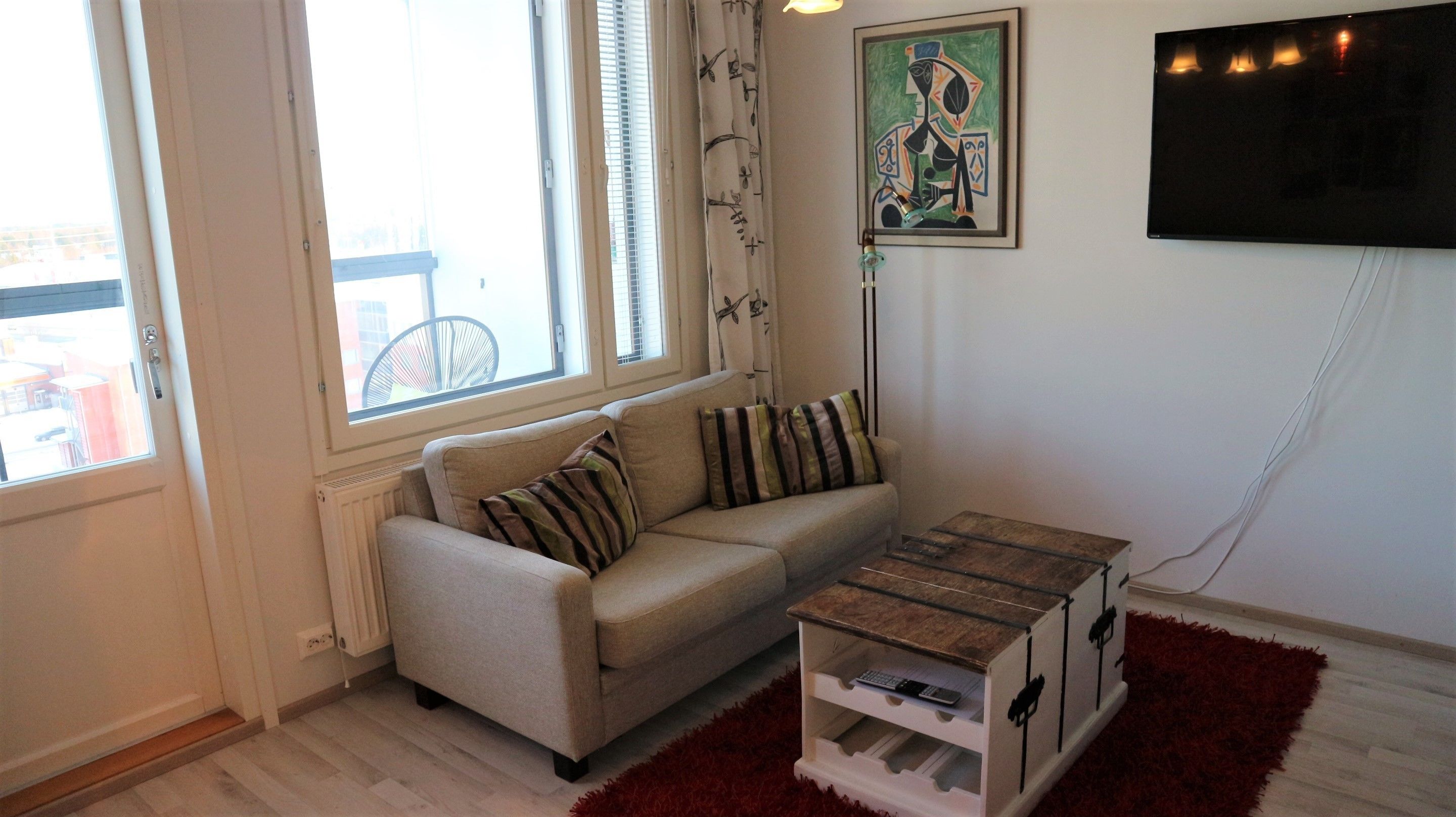 Een woonkamer met een bank, een klein houten bijzettafeltje, een rood vloerkleed, een schilderij aan de muur en een aan de muur bevestigde televisie.