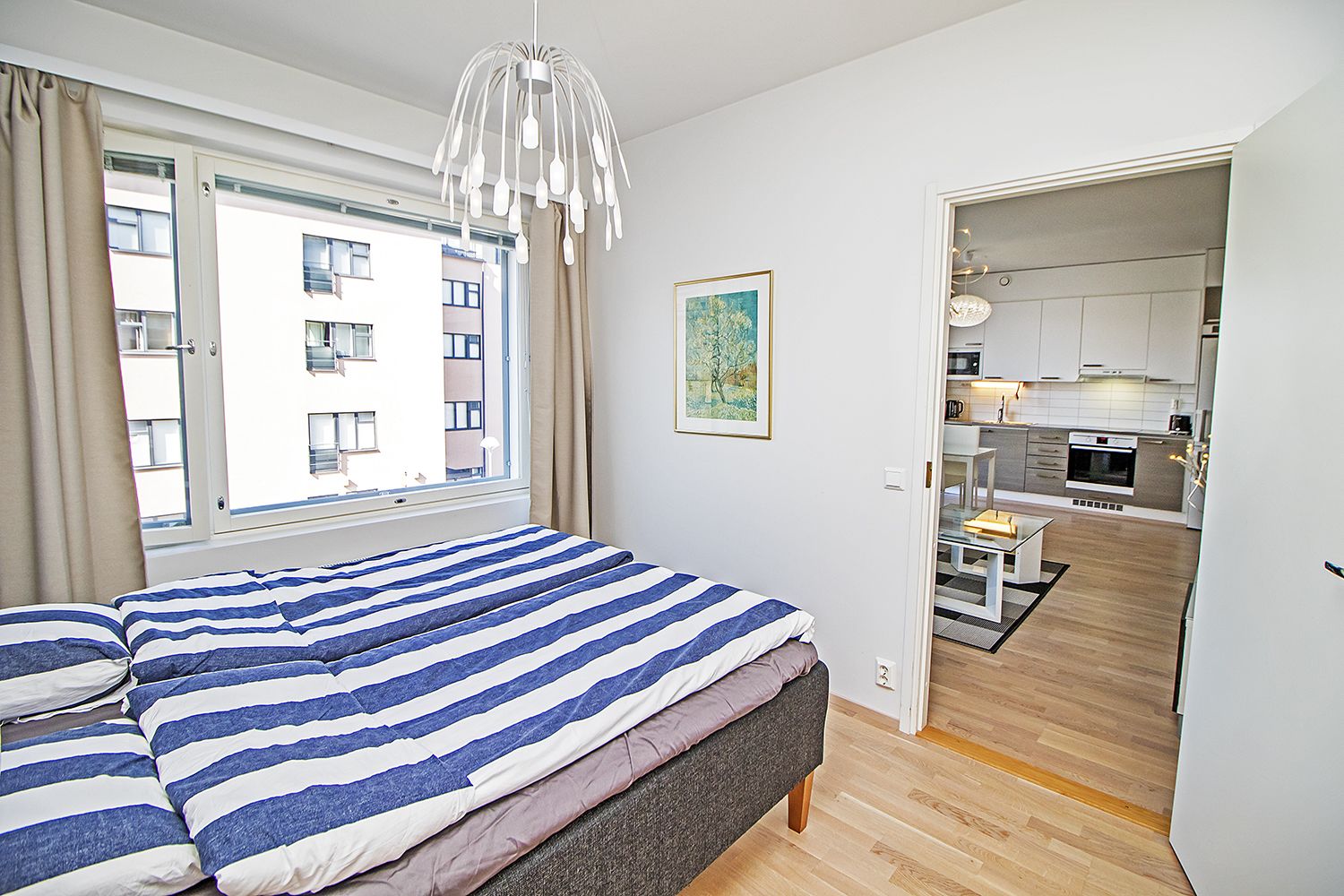 Une chambre à coucher avec un lit, une fenêtre avec des rideaux, un tableau au mur et une vue sur la cuisine par une porte ouverte.