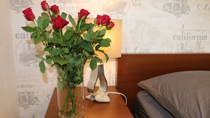 A clear glass vase with red roses on a wooden bedside table next to a bed with a white pillow and a lamp.