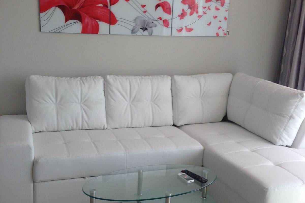 Een moderne woonkamer met een witte leren modulaire bank, een glazen salontafel en een bloemig kunstwerk aan de wand.