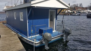 Una casa flotante azul amarrada en un muelle, con una pequeña terraza, una puerta y una ventana.