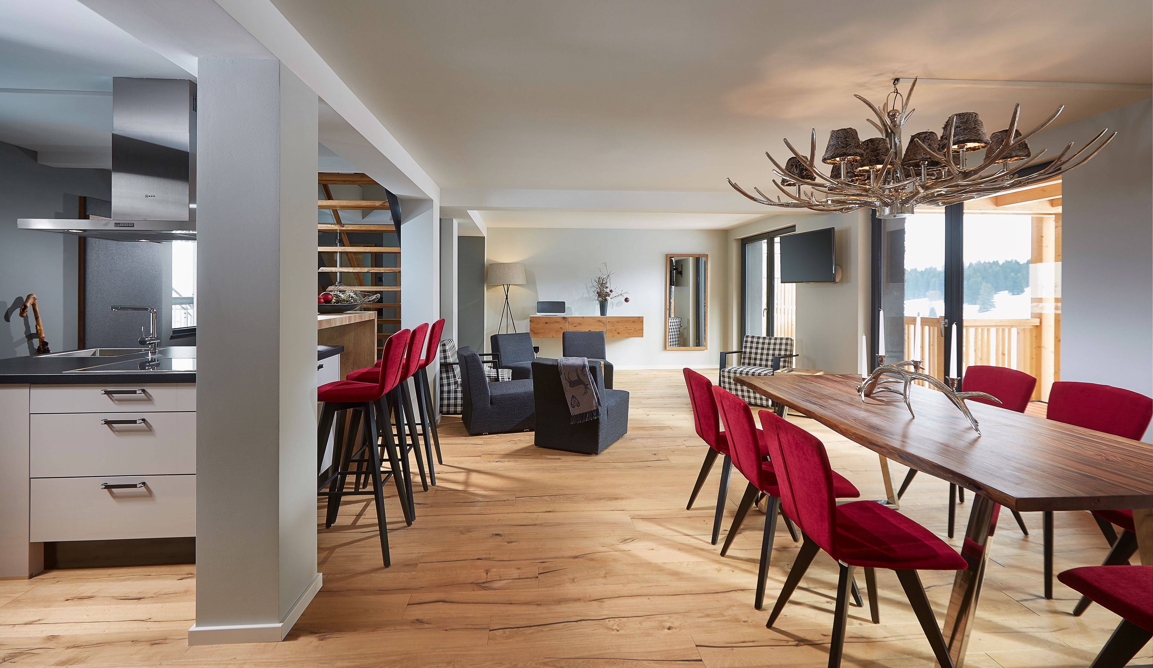 A modern kitchen with a built-in stove, a dining area with a wooden table and red chairs, and a view of a snowy landscape through large windows.
