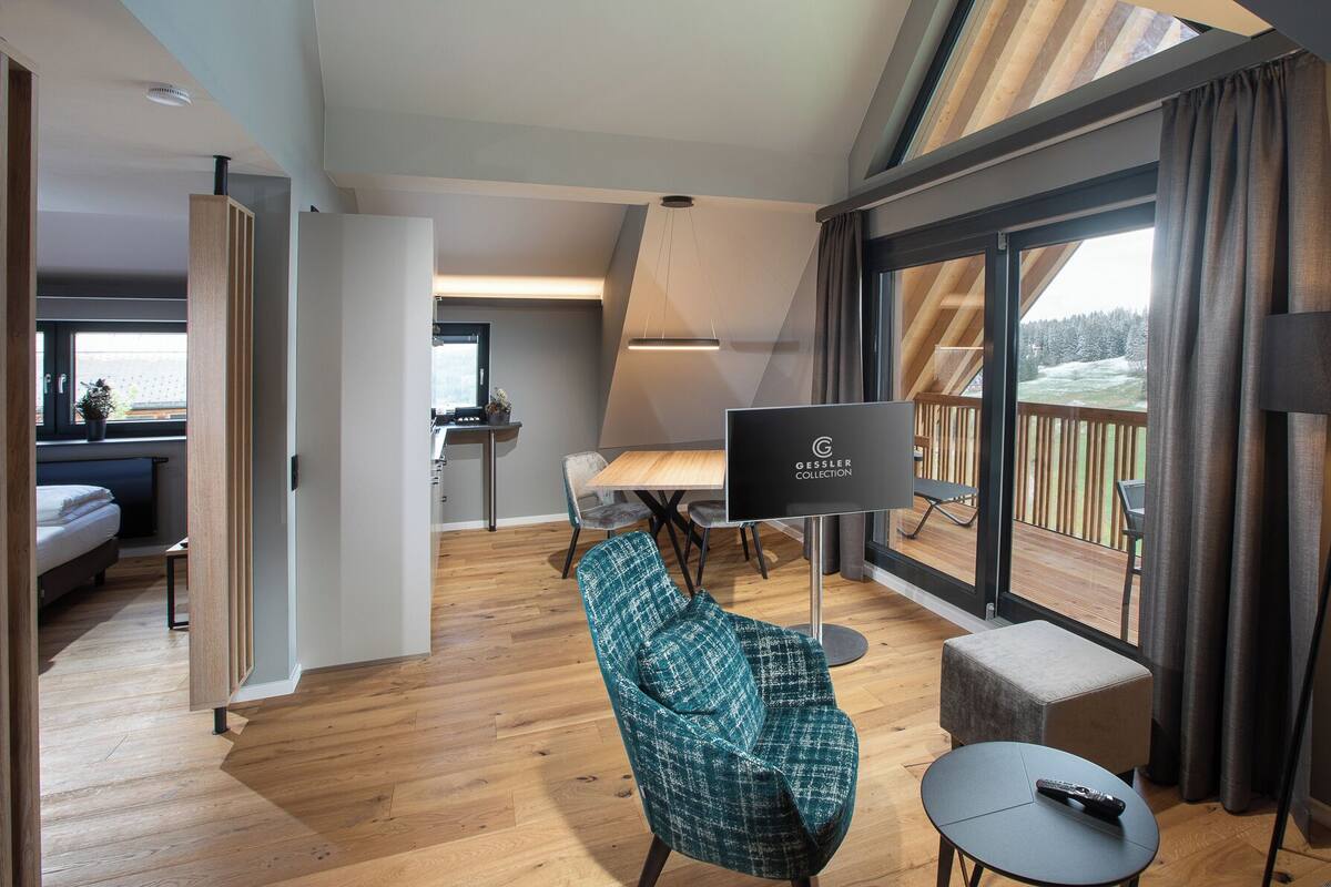 A modern hotel room with a wooden floor, a large window with a view of a snowy landscape, a cozy seating area with a teal chair and ottoman, and a dining area with a table and chairs.