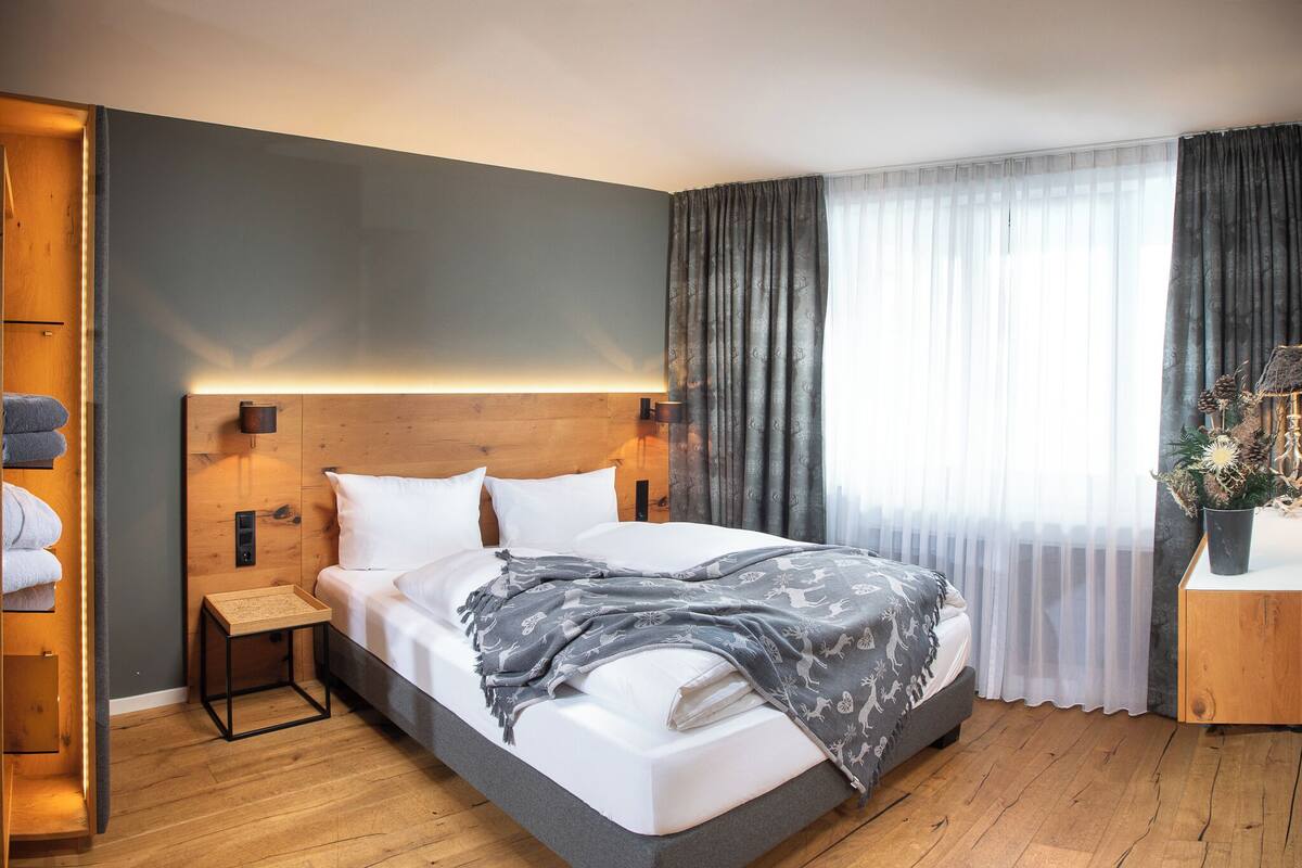 A modern hotel room with a large bed, a wooden headboard, a bedside table, and a nightstand. There is a window with sheer curtains and a dark drape.