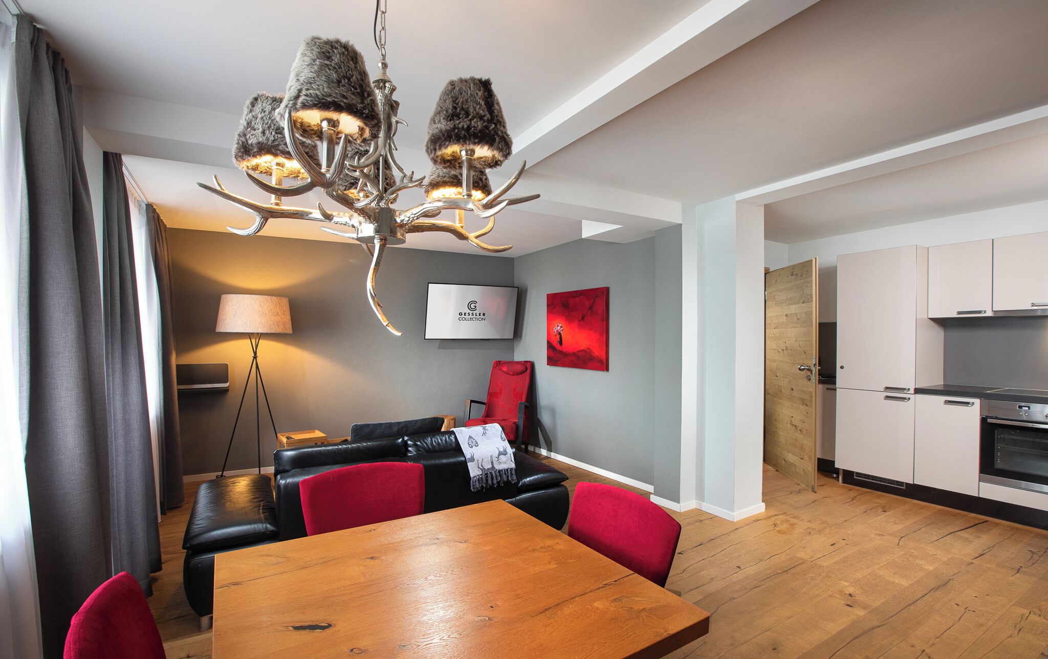 A modern living room with a dining table, a sofa, and a red chair. There is a chandelier with antler decorations, a TV, and a painting on the wall.