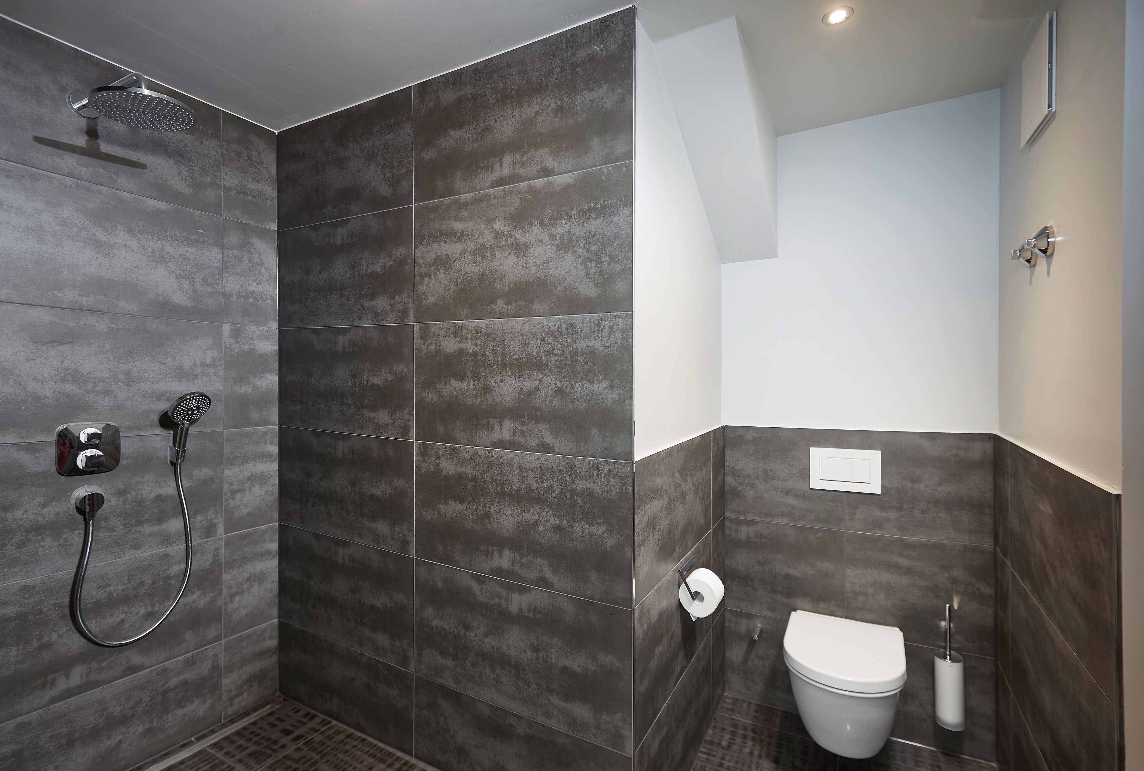 A modern bathroom with dark tiles, a shower, a toilet, and a bidet.