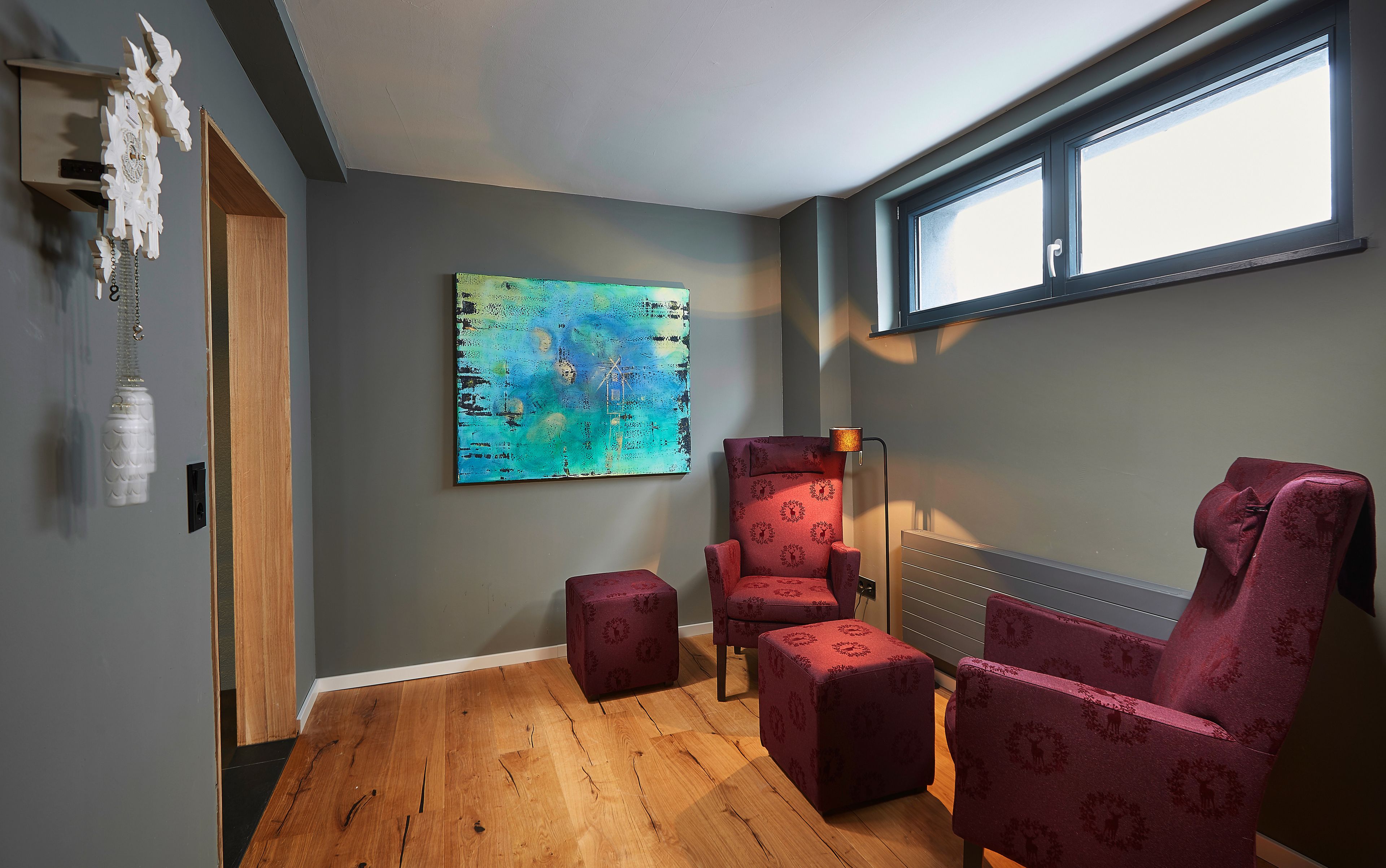 Una habitación con suelo de madera, un sillón rojo estampado, un otoman a juego, un cuadro abstracto de colores y una ventana con vistas.