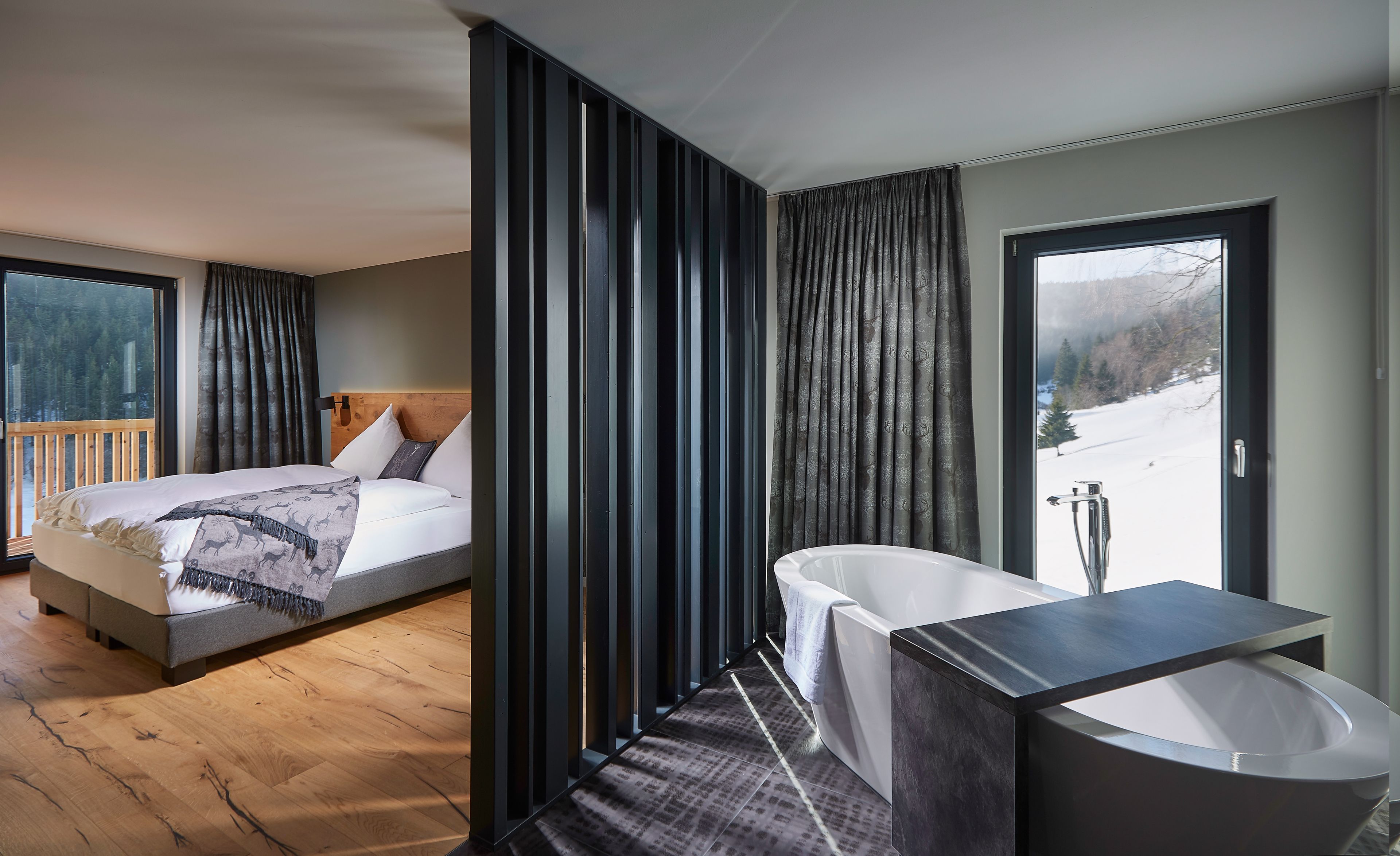 Ein modernes Hotelzimmer mit Holzboden, einem Bett mit Kopfteil, einem Badezimmer mit Badewanne und Blick auf eine verschneite Landschaft.