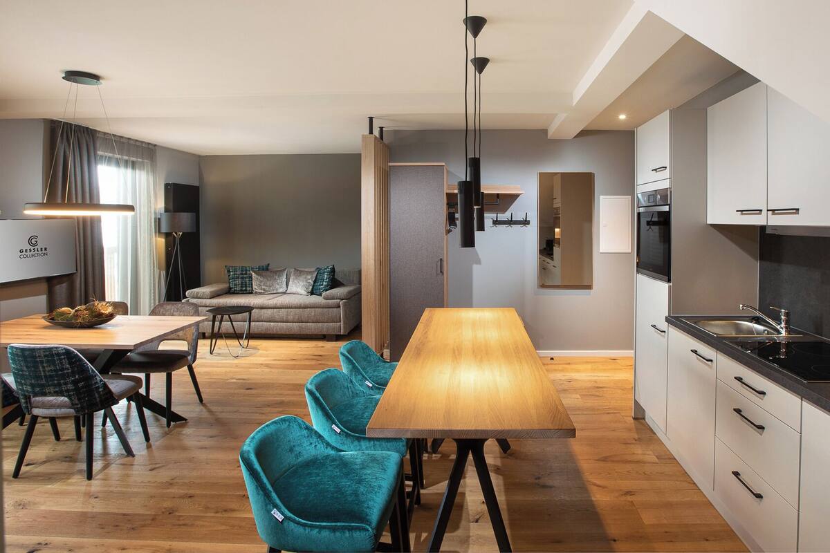 A modern kitchen with a central island, a dining area with a wooden table and teal chairs, and a living space with a sofa and a TV.