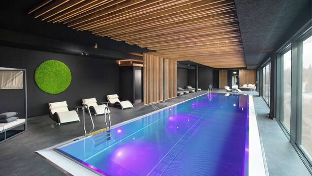 A modern indoor swimming pool with a wooden ceiling, lounge chairs, and a green circular wall decoration. - Black Forest Lodge (Feldberg)