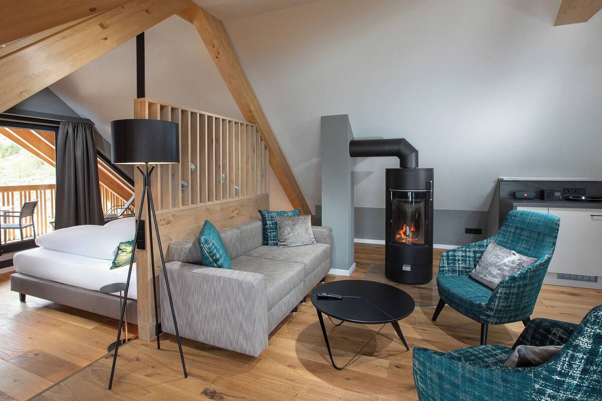 A modern loft bedroom with a large window, a sofa, a floor lamp, and a kitchen area with a stove.