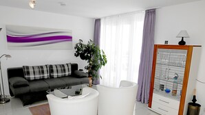 A modern living room with a sofa, two white armchairs, a glass coffee table, a potted plant, a glass cabinet with decorative items, and a lamp. - Ferienwohnung im Atelierhaus (Meersburg)