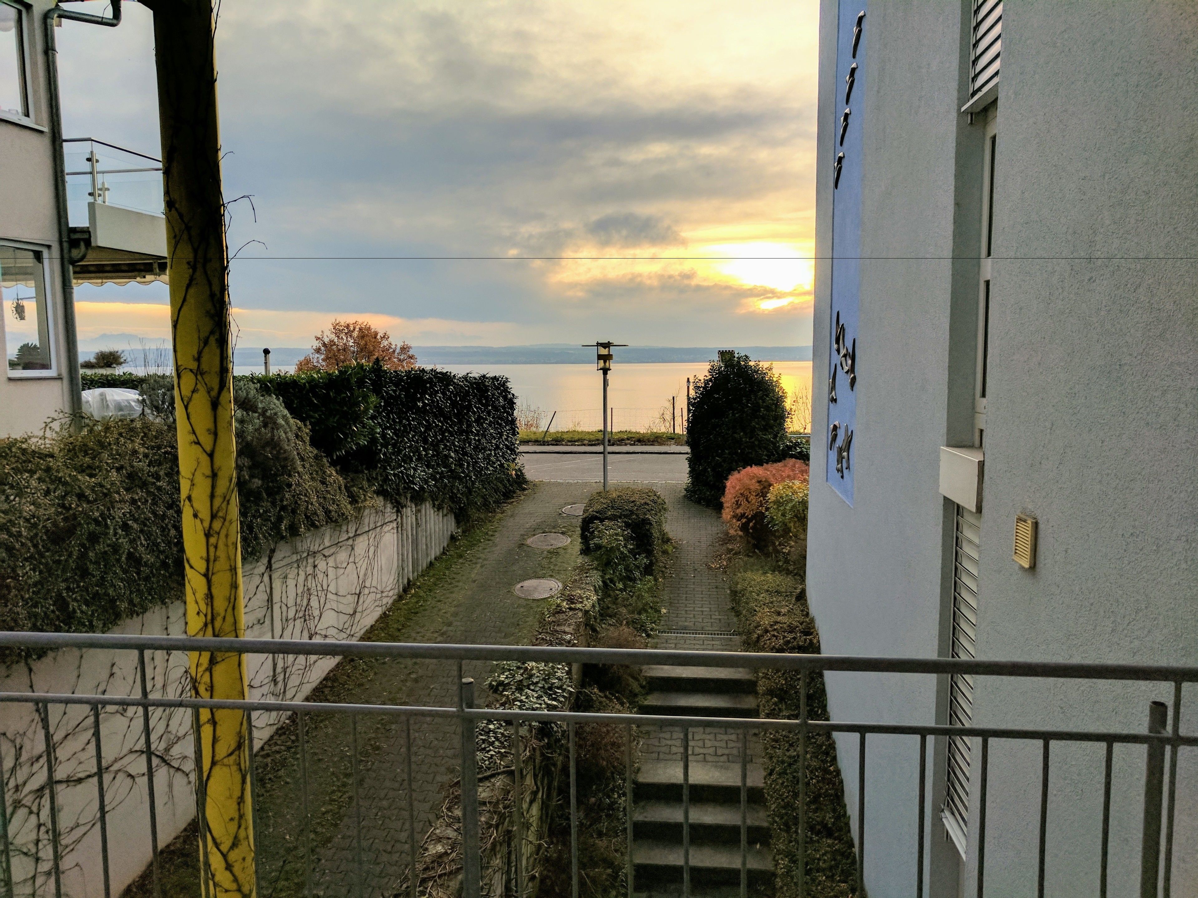 Udsigt fra en balkon over et stiens forløb ned til vandet, med en bygning til højre og en gul pæl til venstre.