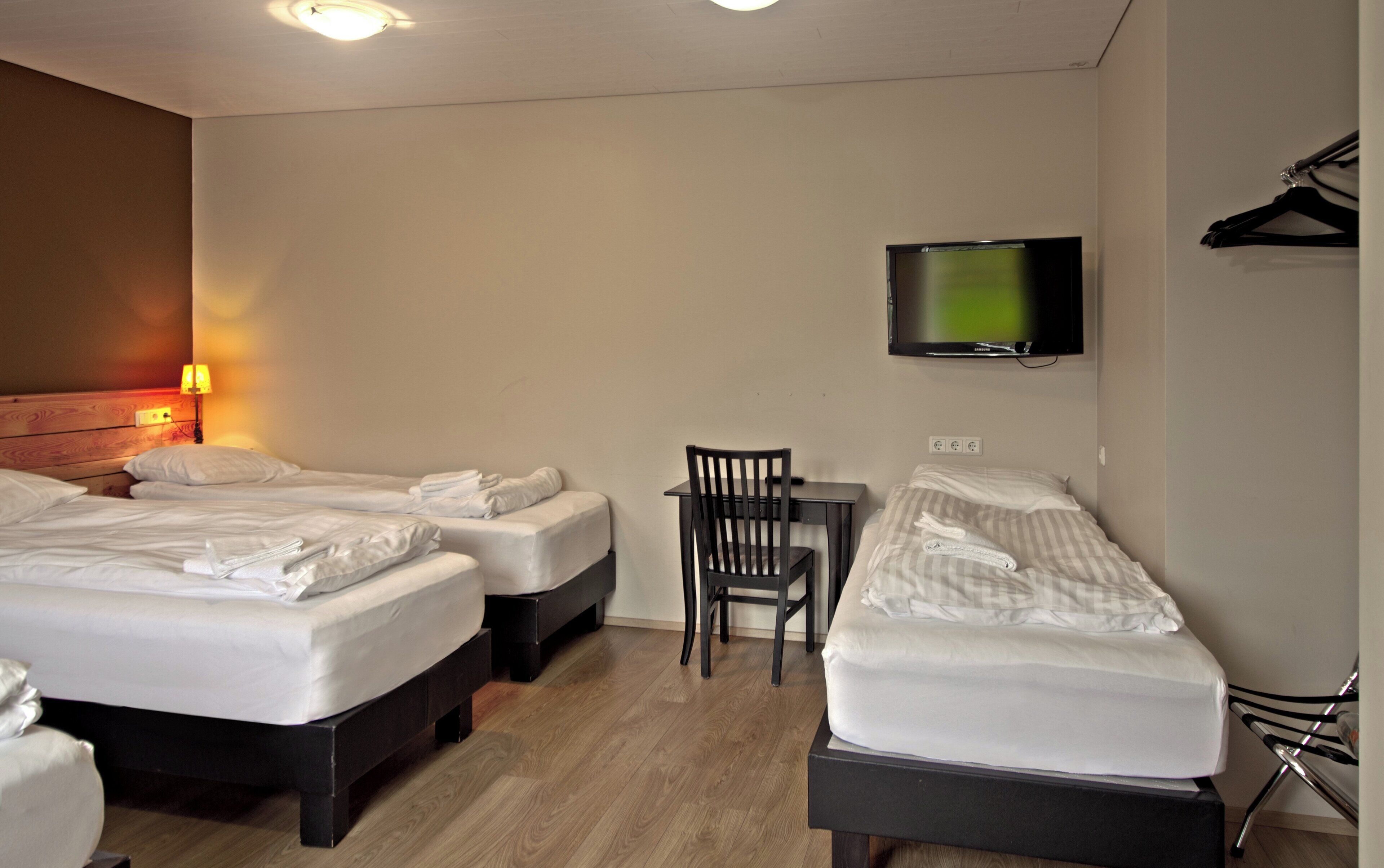 A hotel room with three beds, a desk, a chair, a TV, and a coat rack.
