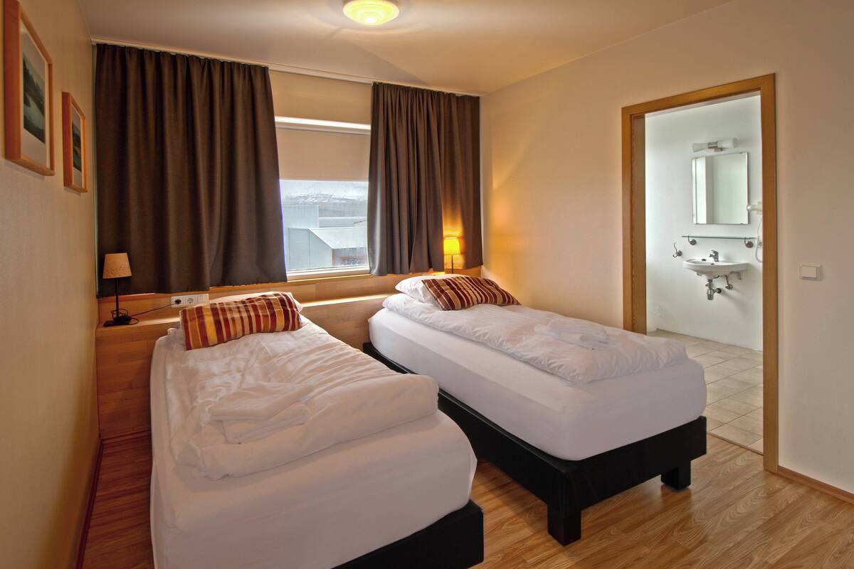 A hotel room with two single beds, a window with curtains, a lamp, and a view of the outside.
