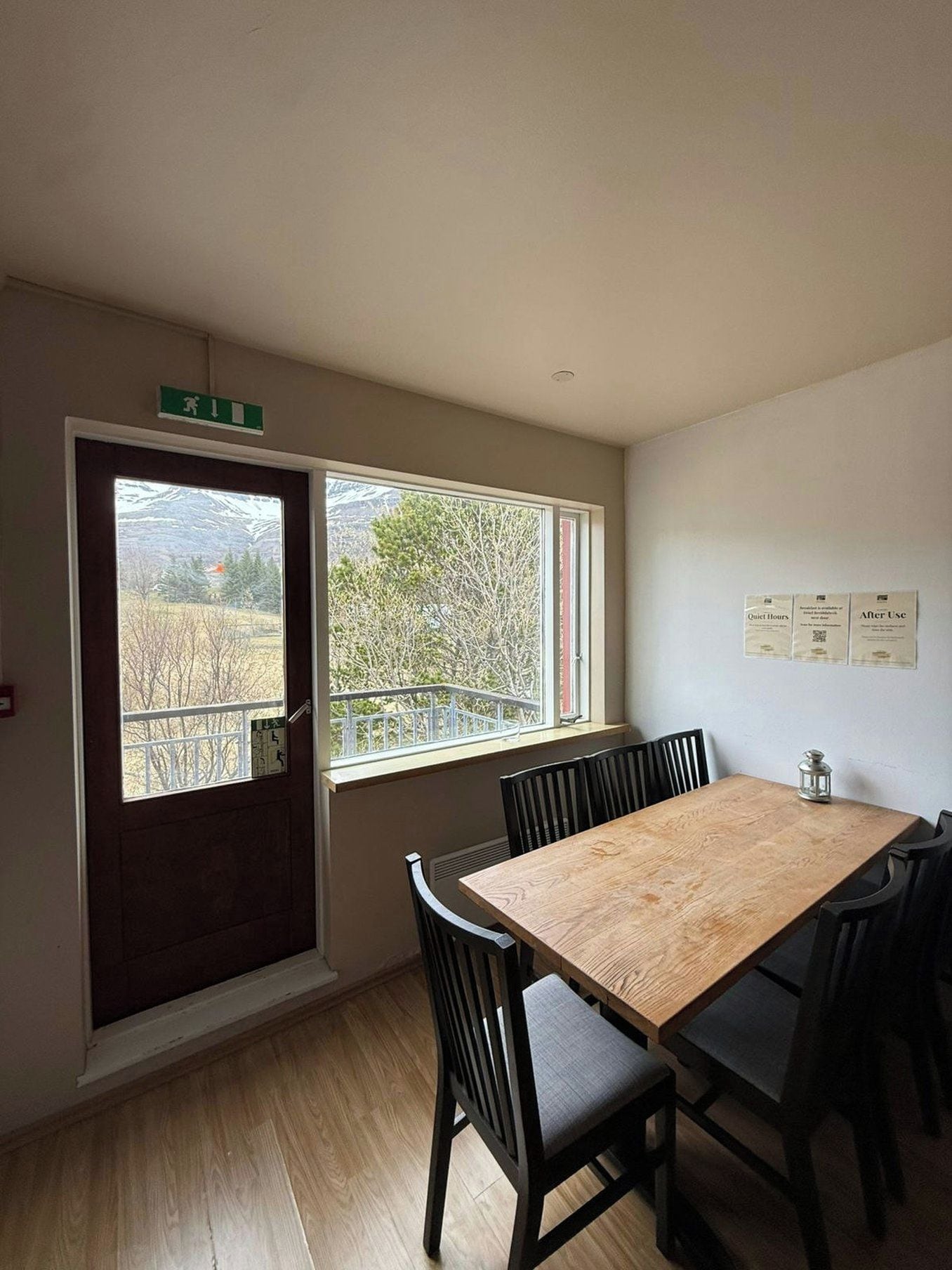 A room with a wooden table and chairs, a window with a view of trees, and a door with an emergency exit sign.