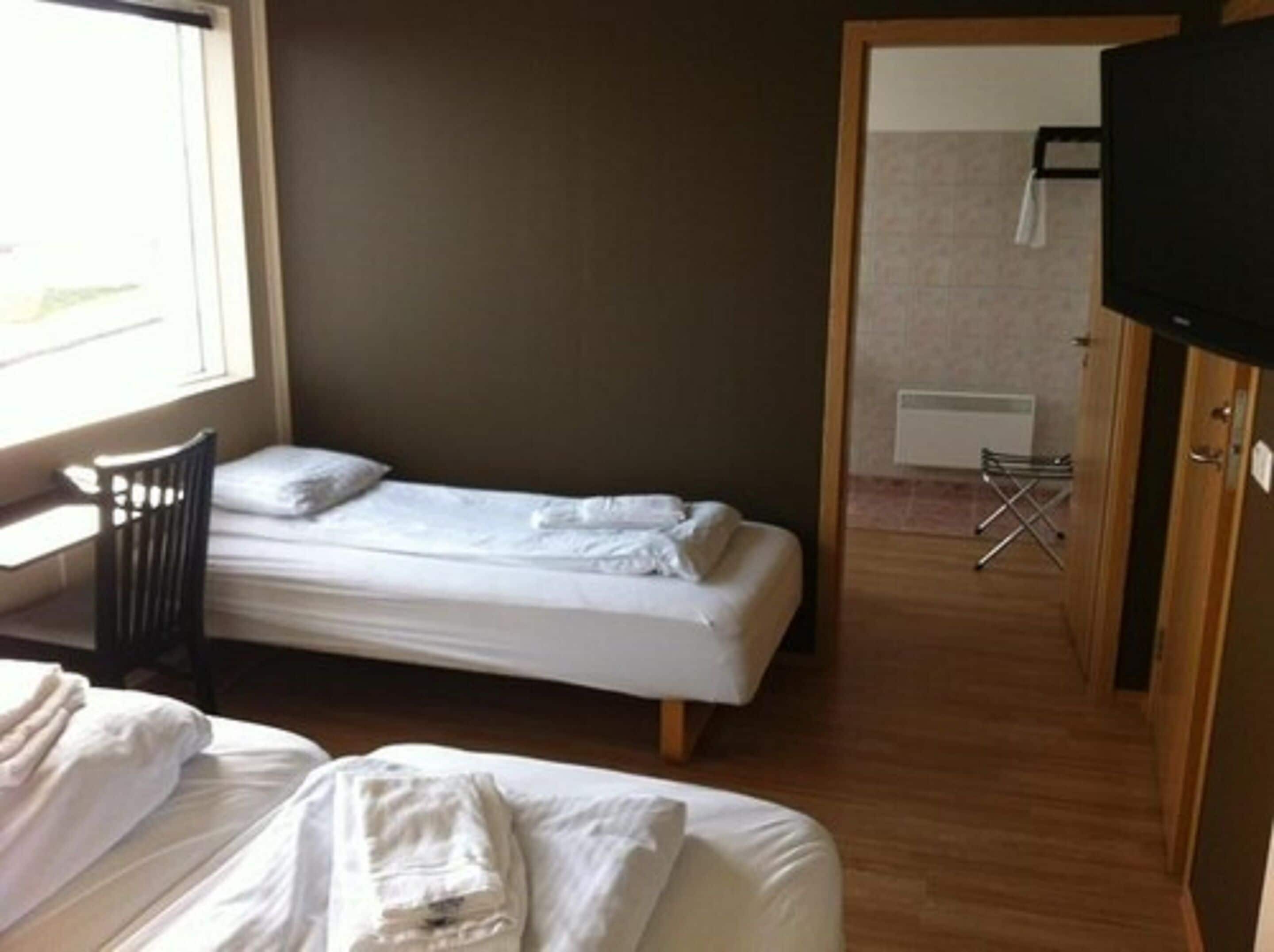 Una cama individual con estructura de madera, una pequeña silla y un televisor sobre un mueble de madera.