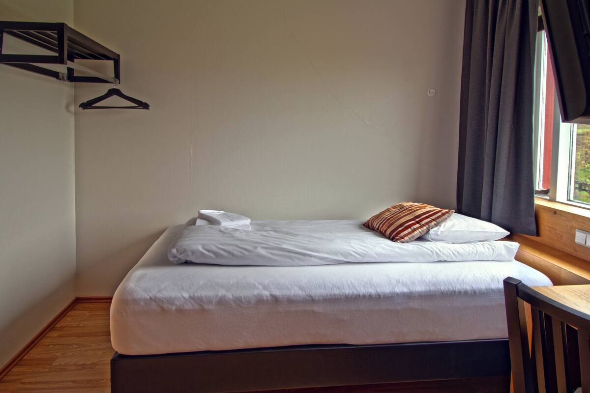 A single bed with white linens, a wooden headboard, and a window with curtains.